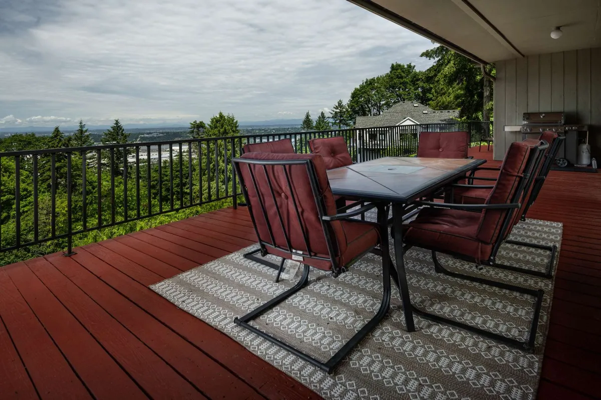 An exterior deck scene with an outdoor dining set, surrounded by lush greenery and panoramic vistas of the river, mountains, and city. The space is set up for both relaxing and entertaining, featuring ample seating and an inviting atmosphere for al fresco gatherings.