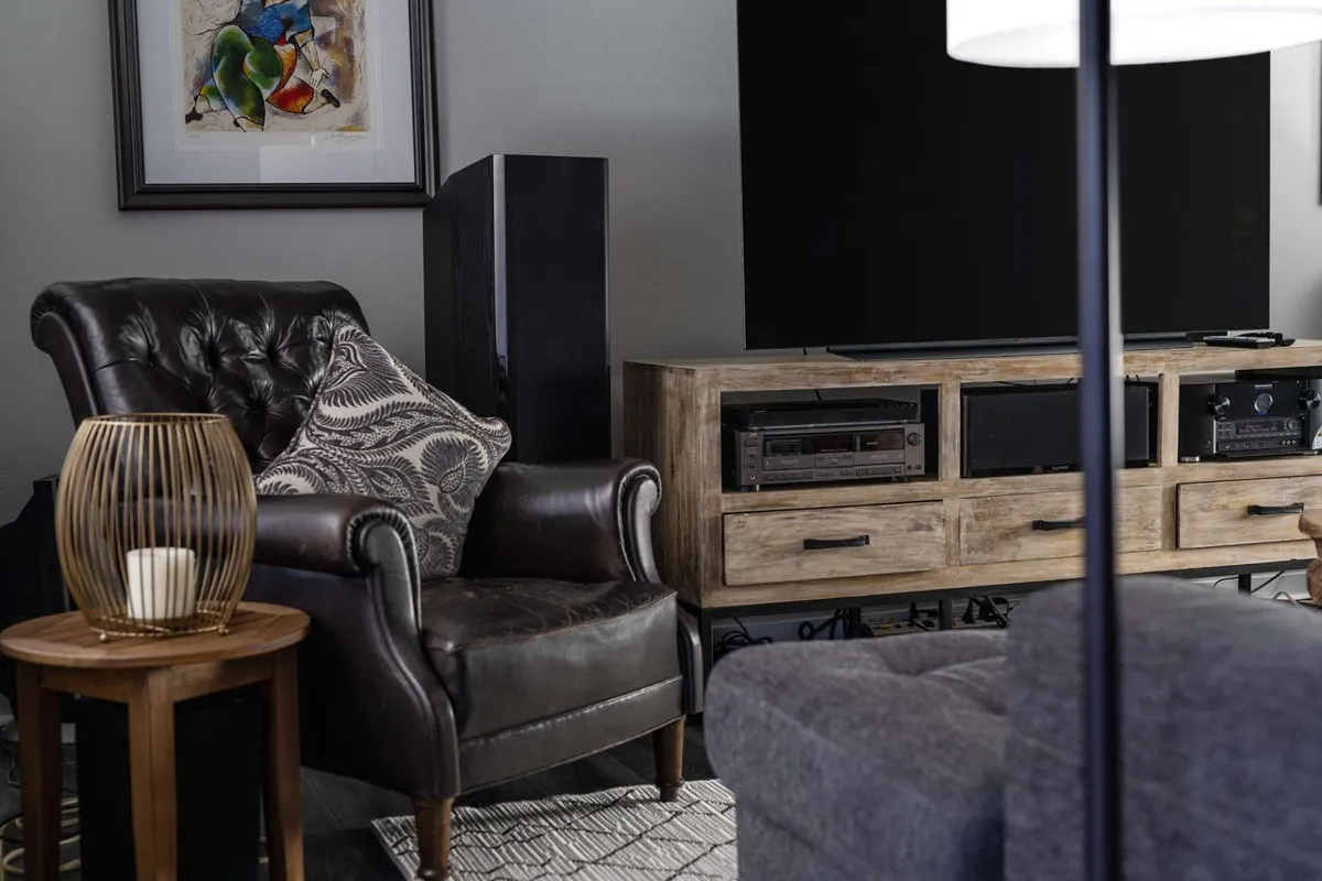 This image captures a cozy living space featuring a leather armchair positioned beside a wooden media console, softly illuminated by a decorative lamp. Framed artwork adds a touch of sophistication, while the setting is staged for relaxing or entertaining guests.