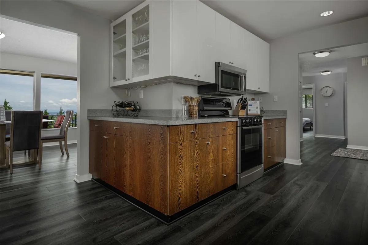 A contemporary interior showing a modern kitchen with sleek cabinetry and stainless steel appliances. The open layout provides a clear view to a dining area set near large windows, allowing in ample light and offering glimpses of the scenic outdoors beyond.