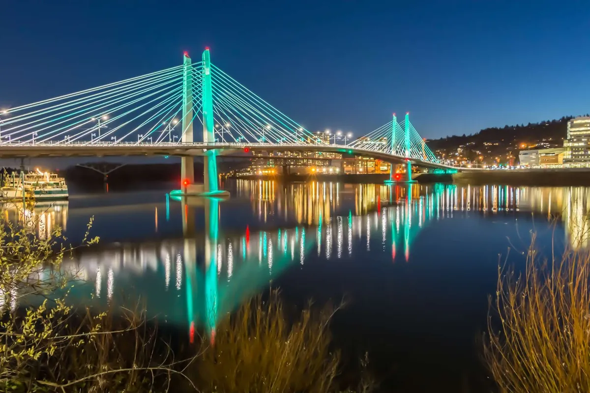 A dynamic evening scene of a bridge with illuminated cables reflected in the river, with city lights sparkling in the background. The image suggests lively riverfront activities, nighttime city exploration, and the appeal of Portland’s vibrant urban life.