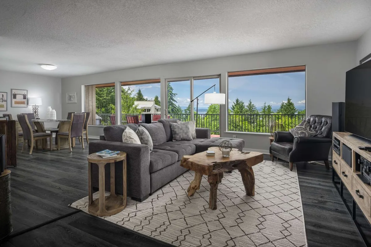 A spacious, light-filled living area featuring a modern gray sectional sofa and a wooden coffee table, surrounded by floor-to-ceiling windows that offer sweeping hillside views. The open layout seamlessly connects the cozy lounge, inviting dining space, and entertainment center, all framed by natural light and warm, welcoming decor.