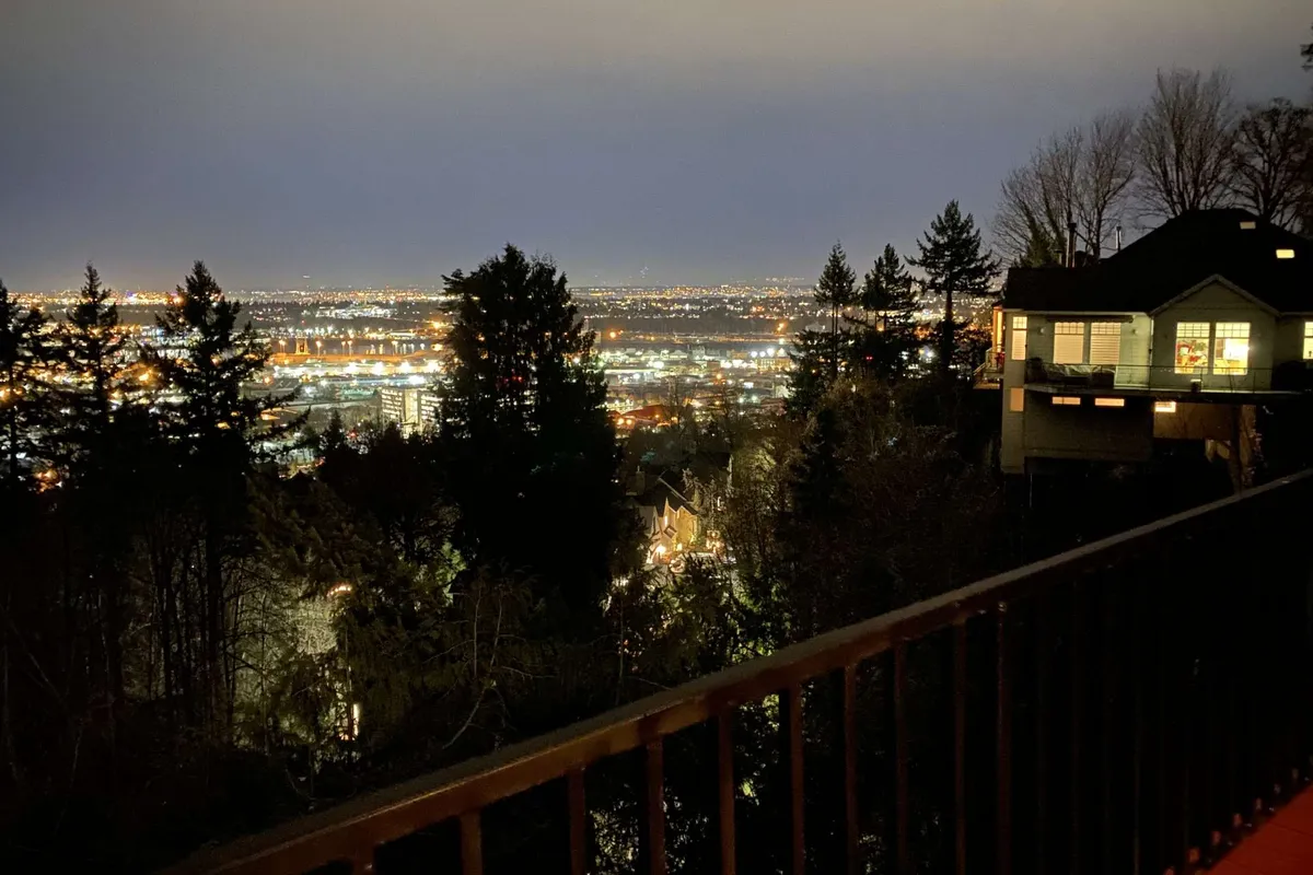 This image shows the city lights sparkling at dusk, with a distant skyline, surrounding trees, and the upper part of a house in the foreground. The mood is tranquil, capturing a peaceful evening and highlighting the elevated vantage point perfect for twilight gatherings or quiet contemplation.