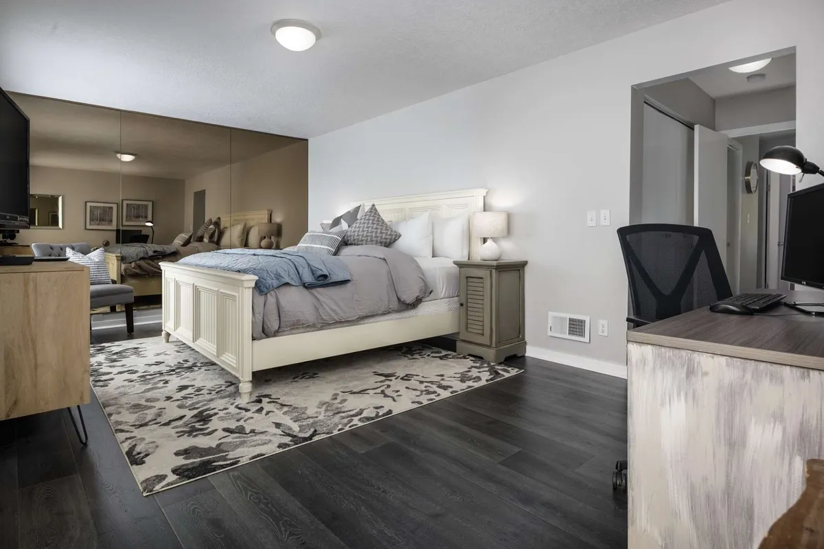 A cozy bedroom featuring a plush, inviting bed with soft linens and decorative pillows. The nightstand with a lamp offers gentle lighting, and a desk with a computer provides a versatile space for reading or working. A mirror reflects the peacefulness of the room, tying together the tranquil, comfortable design.
