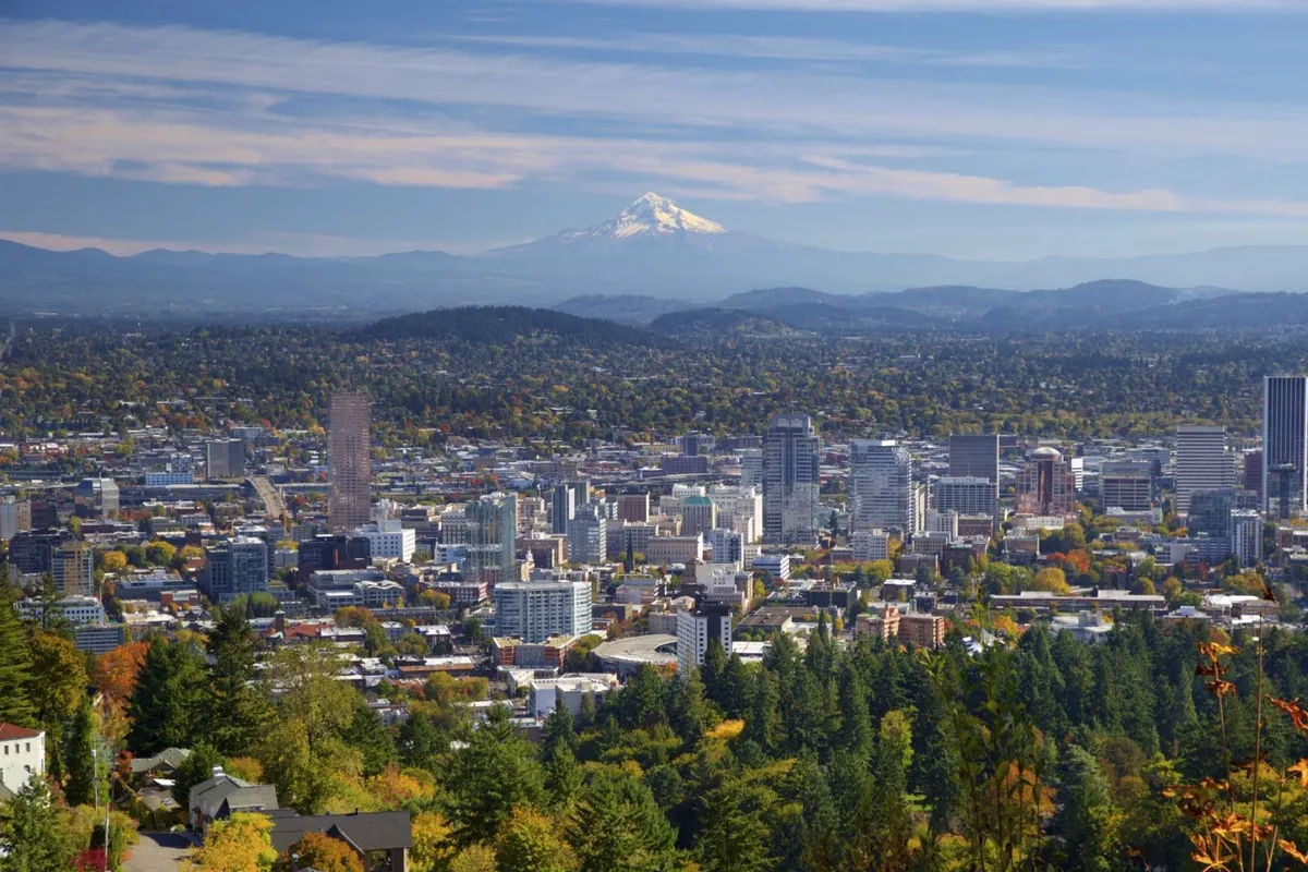 A sweeping view of Portland's downtown skyline with Mount Hood in the distance, framed by a rich line of green trees in the foreground. The image evokes a seamless connection between urban life and natural beauty, showcasing the accessibility of the city’s attractions alongside lush scenery.